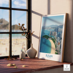 Framed Busan skyline travel poster on window sill with vases, beach view outside window