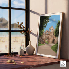 Framed Colchester travel poster on a wooden windowsill with vases and beach view.