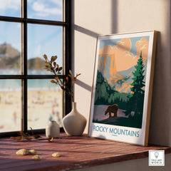 Framed Rocky Mountains Canada travel poster on windowsill with vases and scenic view outside.