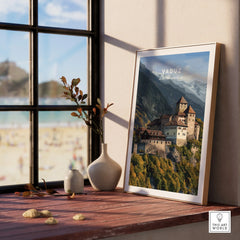 Framed Vaduz castle travel poster on windowsill with vases and mountain landscape print