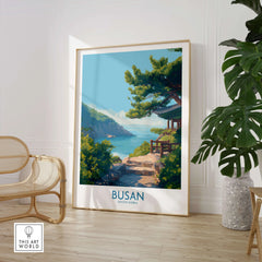 Framed Busan travel poster with coastal landscape, beside chair and large indoor plant