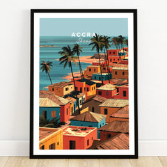 Accra Ghana coastal village travel poster, terracotta rooftops and palms by a turquoise sea, in a black frame on a wooden floor