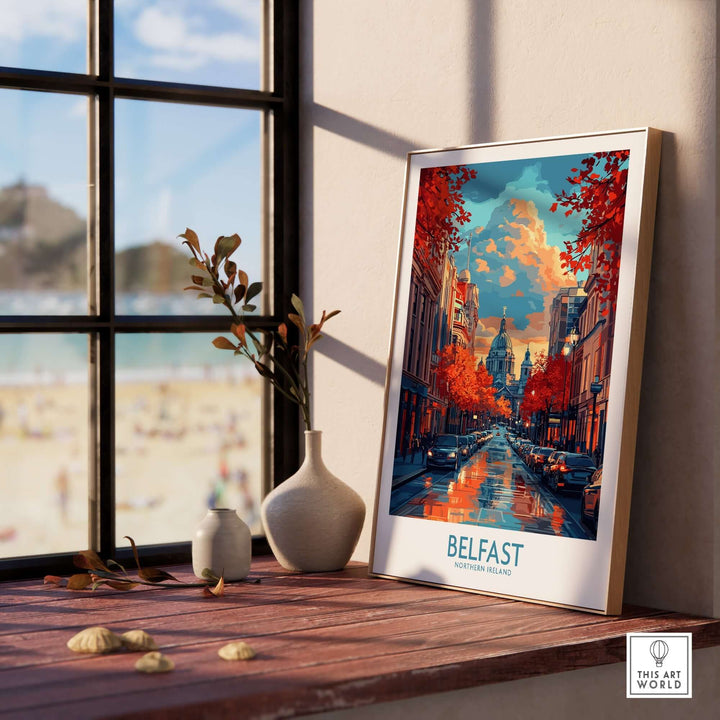 Belfast travel poster showcasing vibrant city scenery with autumn colors, framed on a wooden table by a window.