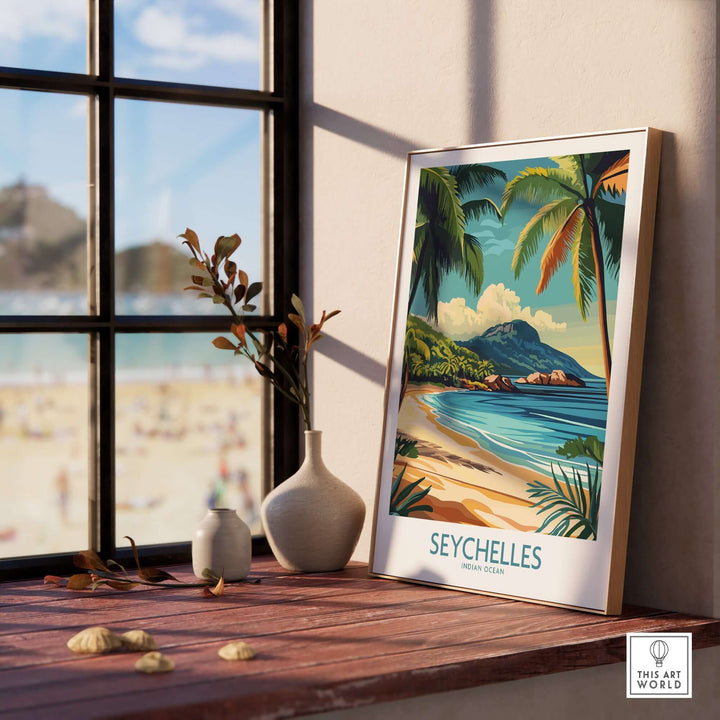 Framed Seychelles beach art print on a windowsill with a view of a sandy beach and palm trees.