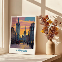 Aberdeen Scotland travel poster with city skyline, red bus, and sunset, displayed on windowsill with dried flowers.