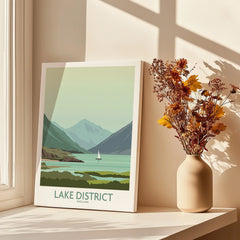 Lake District England travel poster with mountains, lake, and sailboat, styled as wall art beside dried flowers.