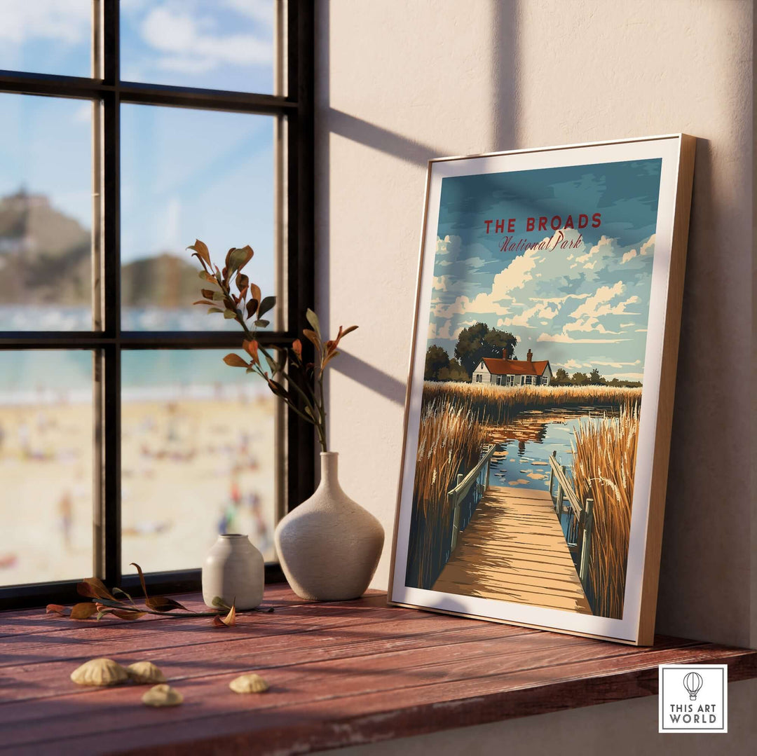 The Broads National Park travel print displayed on a wooden shelf with a sunlit beach view through the window.