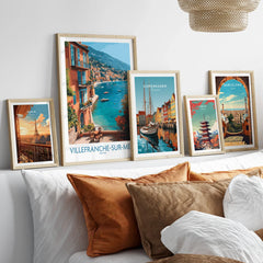 Set of five framed travel art prints featuring Paris, Villefranche-sur-Mer, Copenhagen, Tokyo, and Barcelona above a bed with earth-tone pillows.