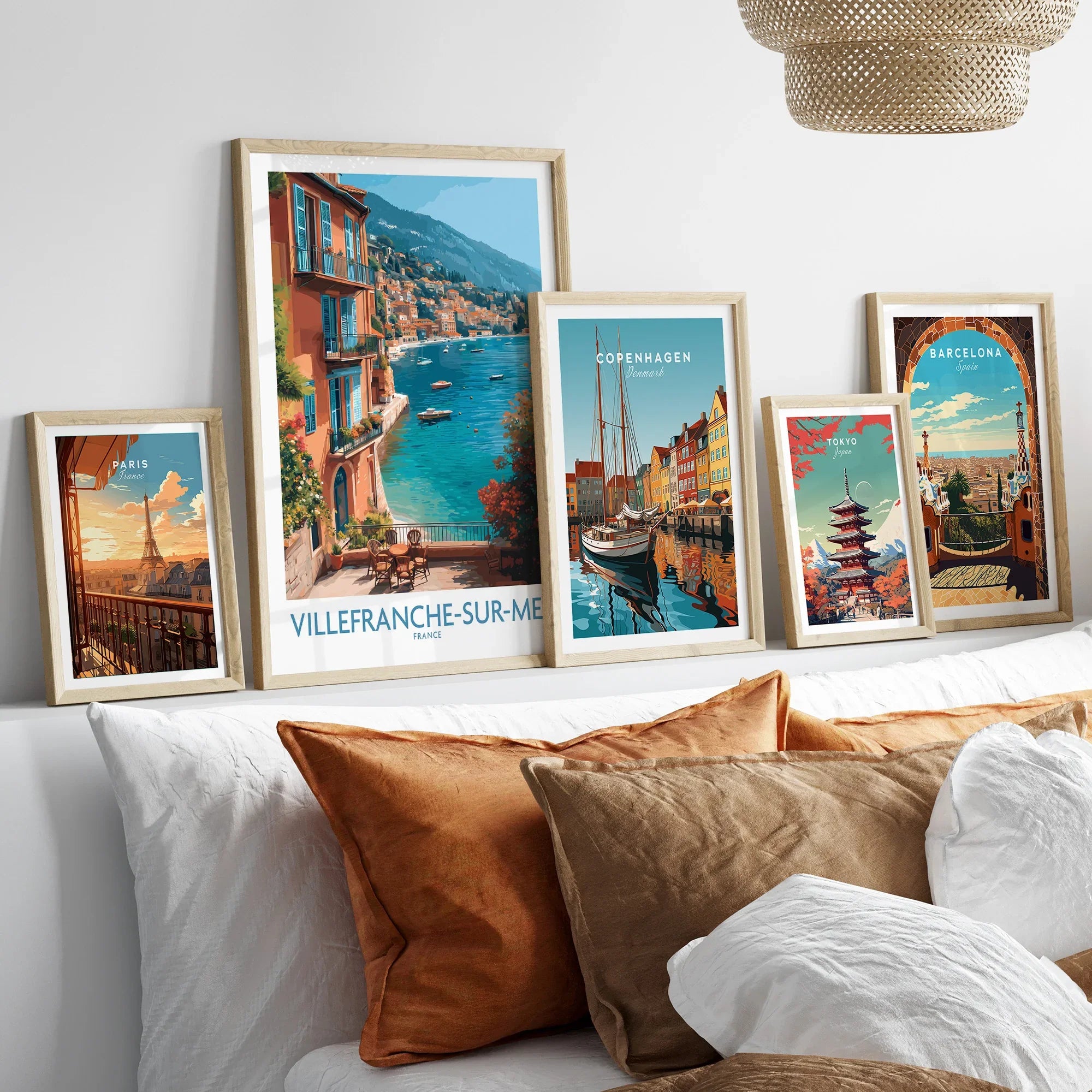 Set of five framed travel art prints featuring Paris, Villefranche-sur-Mer, Copenhagen, Tokyo, and Barcelona above a bed with earth-tone pillows.