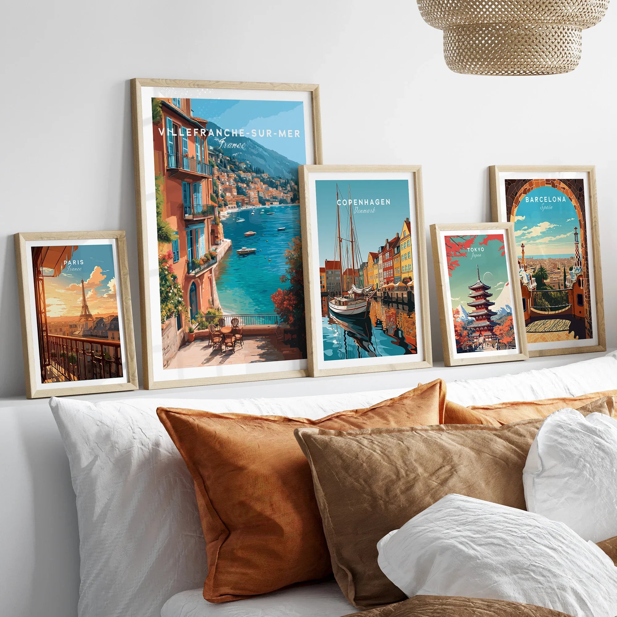 Set of five framed travel art prints featuring Paris, Villefranche-sur-Mer, Copenhagen, Tokyo, and Barcelona above a bed with neutral wood frames.