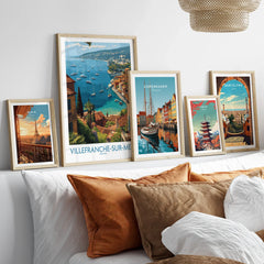 Set of five travel posters in natural wood frames above bed, featuring Paris, Villefranche-sur-Mer, Copenhagen, Tokyo, and Barcelona wall art.