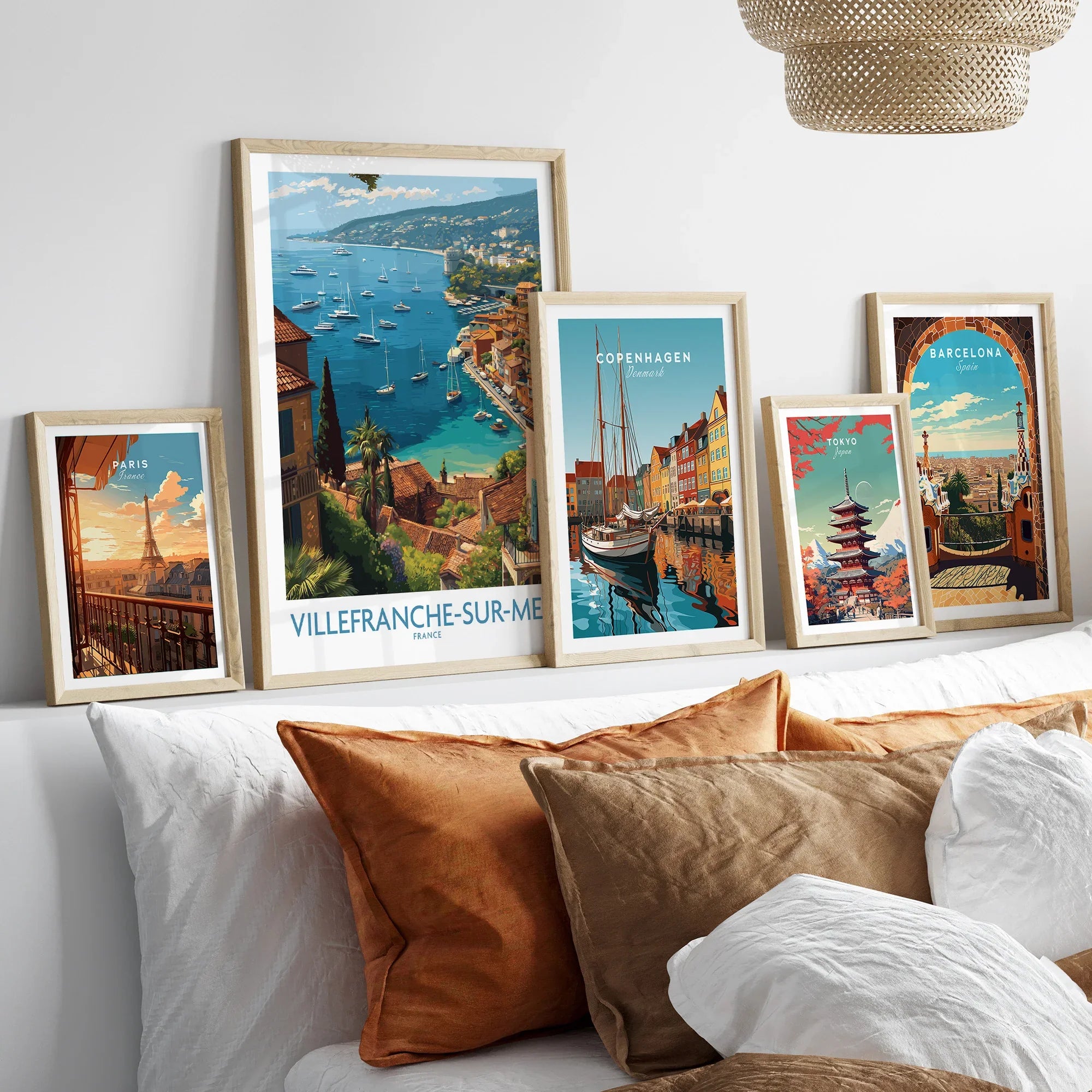 Set of five travel posters in natural wood frames above bed, featuring Paris, Villefranche-sur-Mer, Copenhagen, Tokyo, and Barcelona wall art.