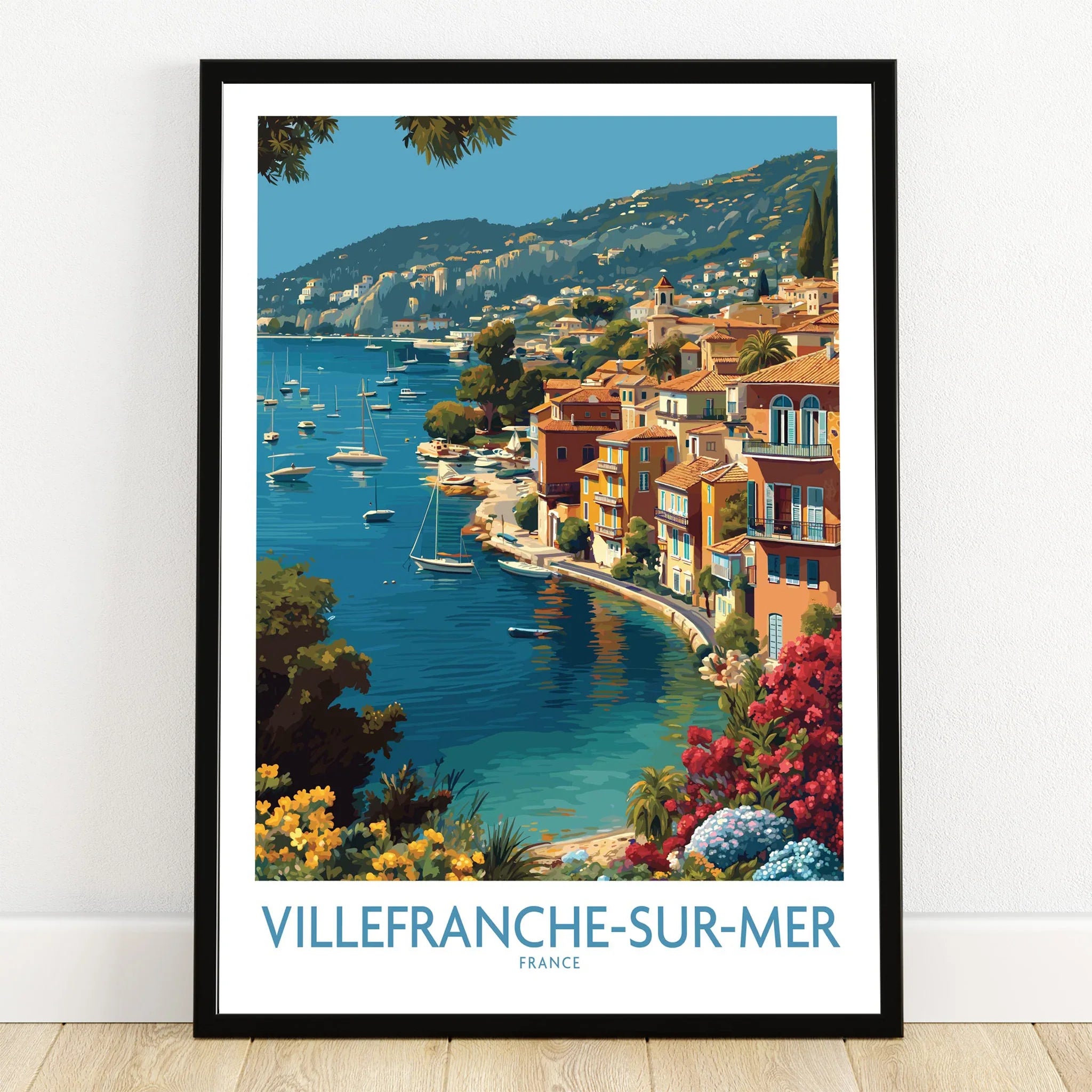 Villefranche-sur-Mer France travel poster art print in black frame on wooden floor, featuring colorful coastal buildings and boats on blue water.