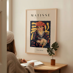 Matisse Art Prints - Woman in Patterned Hat & Dress Poster displayed in a cozy setting with a person reading.