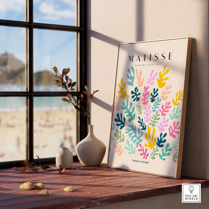 Matisse art print featuring colorful abstract botanical shapes on beige background, displayed on a wooden table by a window.
