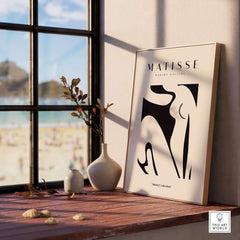 Matisse Art Prints - Black Abstract Nude Figure Poster displayed on a wooden table by a window with beach view.