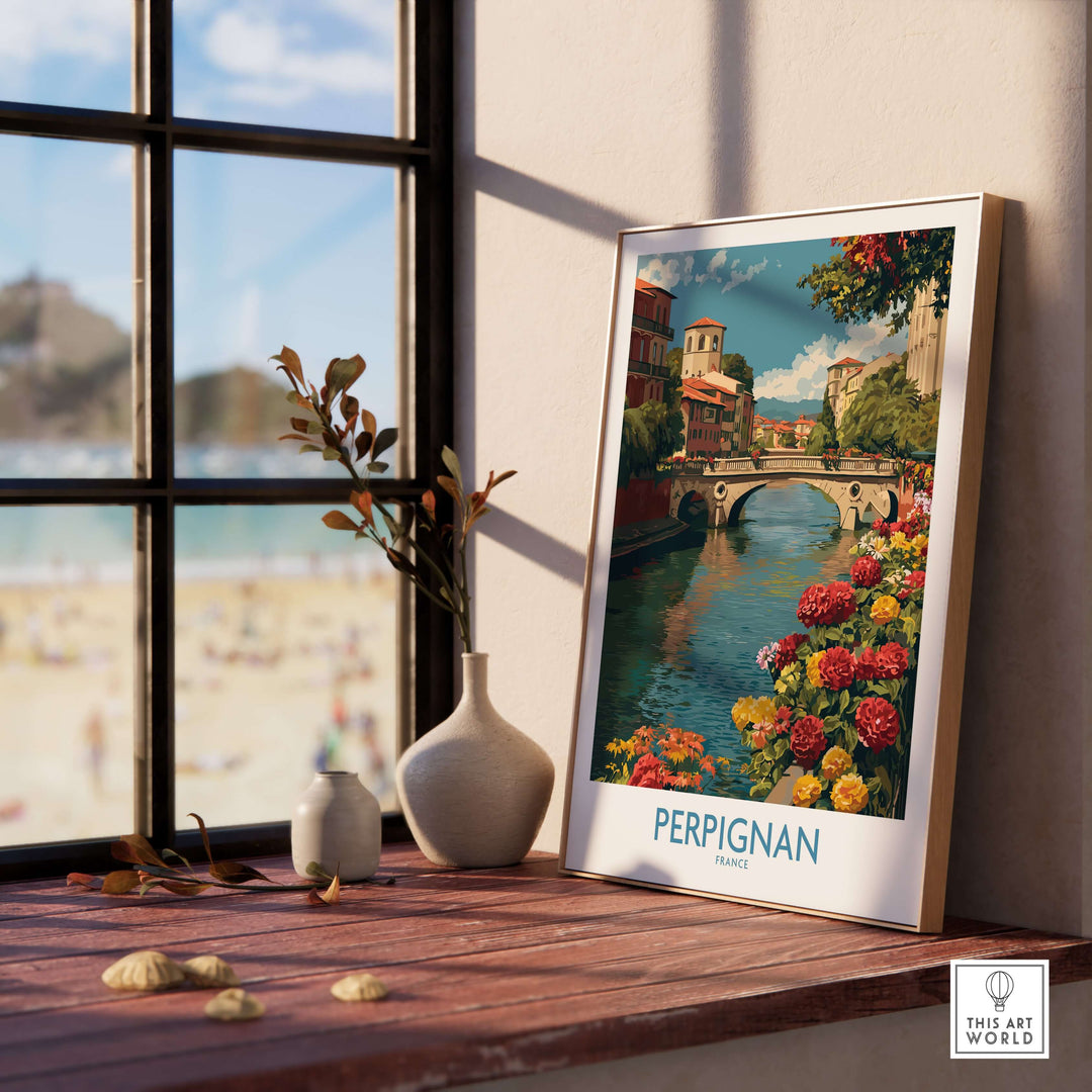 Perpignan River poster in a frame, showcasing a bridge and colorful flowers, displayed indoors near a sunny window.