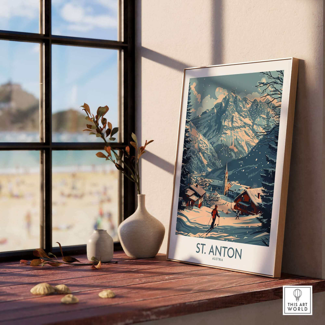 Framed St. Anton ski poster on wooden table by window, depicting snowy Austrian mountains and village.